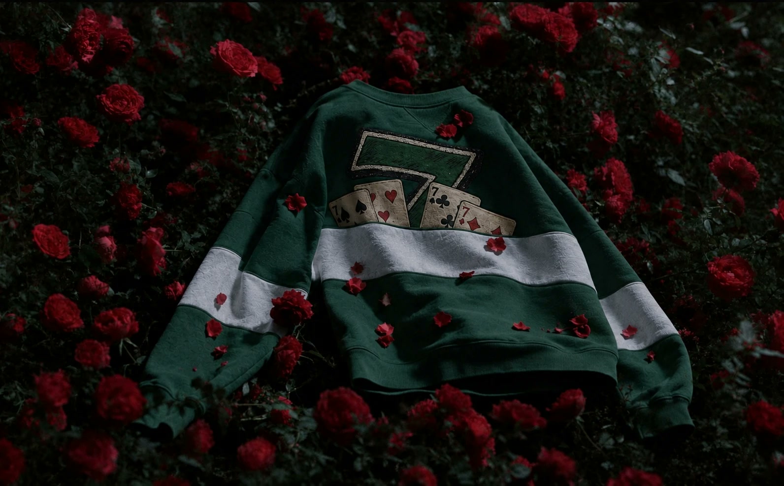 Sudadera verde de HARES caída sobre un campo de rosas rojas de noche, con el siete y las cuatro cartas cosidos en la espalda